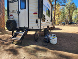A cream-colored rv rental, a Sun Lite Sport, is set up at a sunny forest campsite, powered by a generator and propane tank.