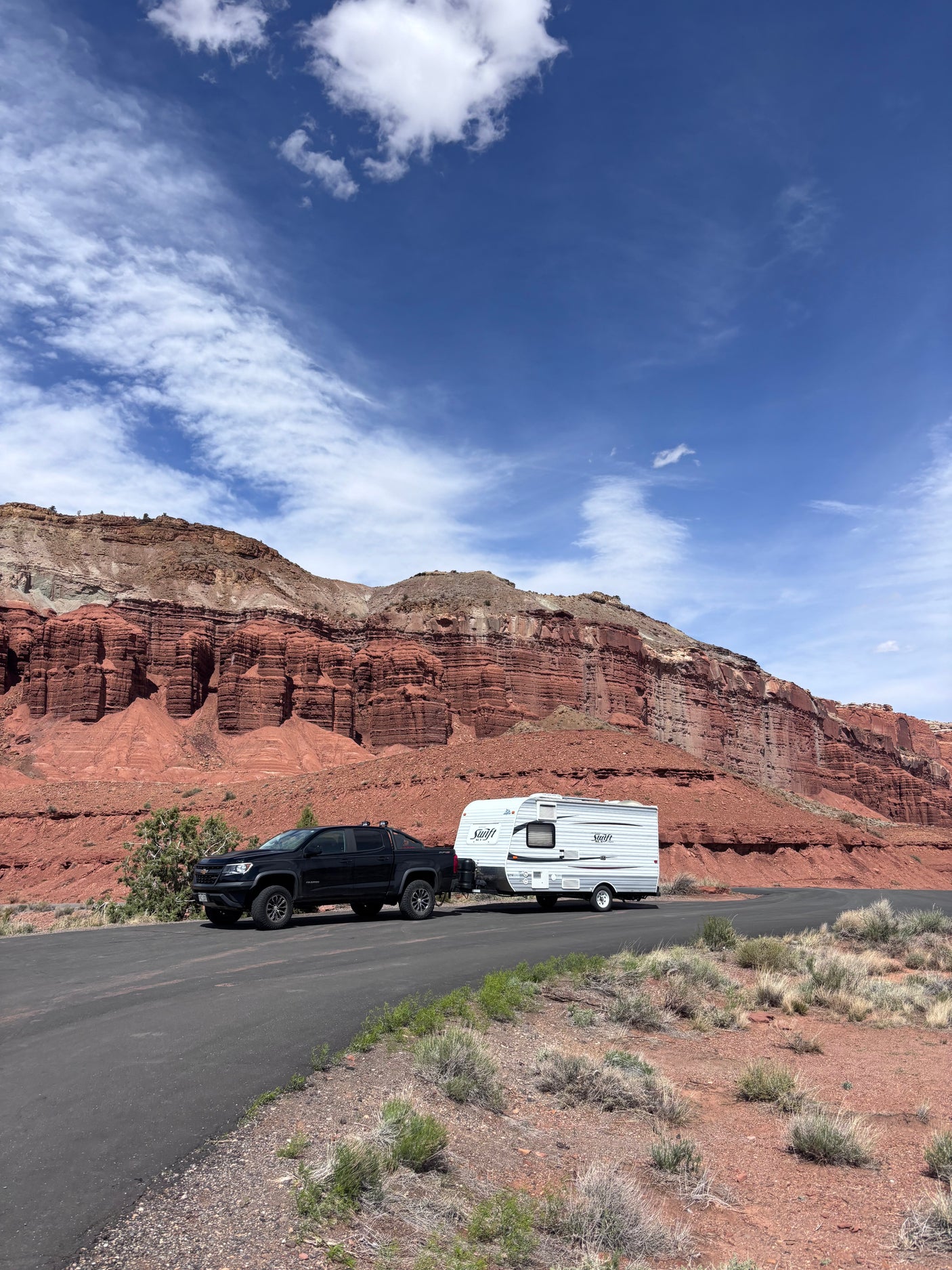 Black pickup truck towing an rv rental travel trailer on a road beside towering red rock cliffs, under a blue, cloudy sky.