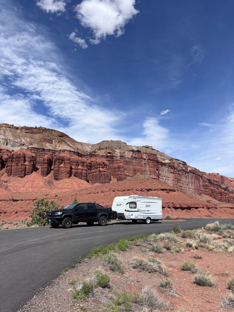 Black pickup truck towing an rv rental travel trailer on a road beside towering red rock cliffs, under a blue, cloudy sky.