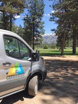 A silver Wandervans RV rental van parked among pine trees with snow-capped mountains & a green field in the distance under a blue sky.