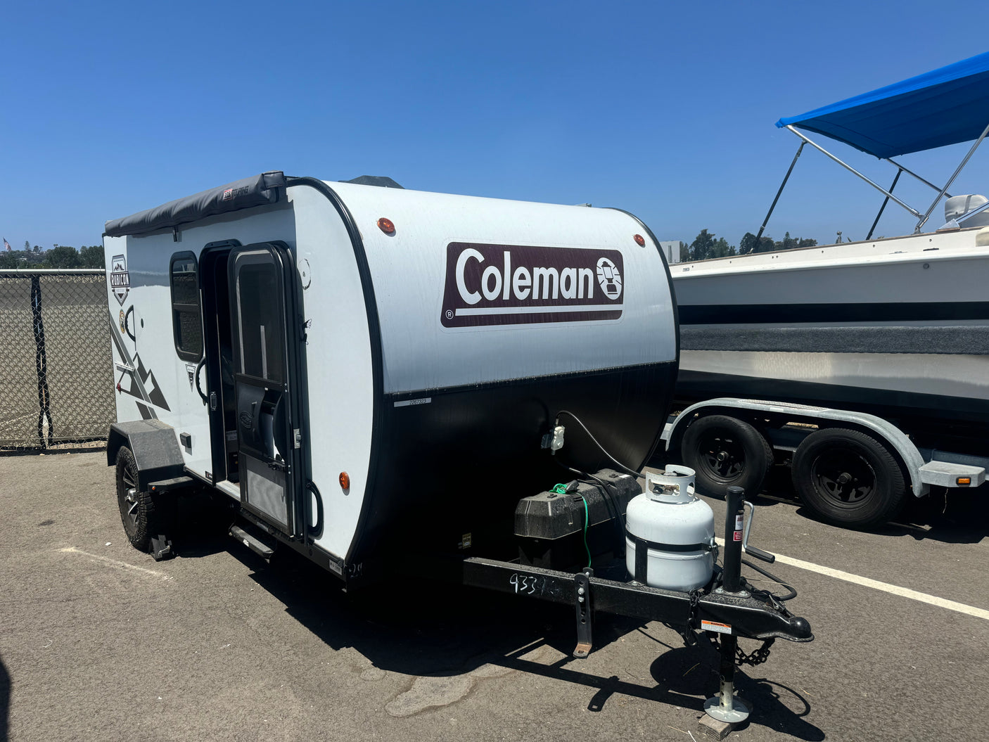 A white and black Coleman camper trailer (RV rental) is shown on asphalt, parked beside a boat. Ready for outdoor adventures.