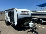 A white and black Coleman camper trailer (RV rental) is shown on asphalt, parked beside a boat. Ready for outdoor adventures.