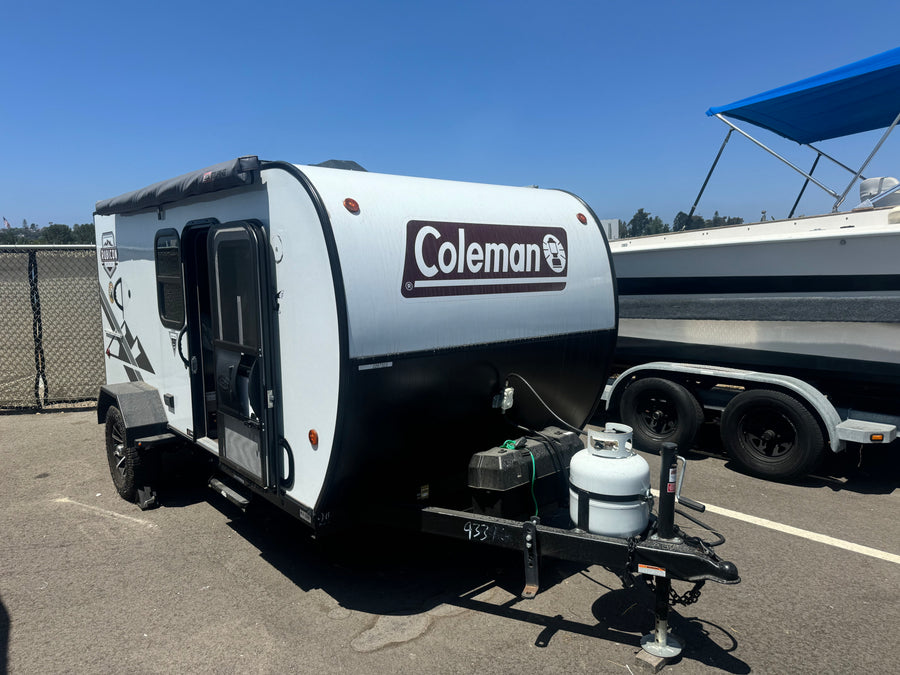A white and black Coleman camper trailer (RV rental) is shown on asphalt, parked beside a boat. Ready for outdoor adventures.