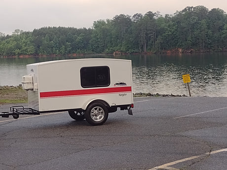 RV rental: A white compact camper trailer, featuring a red stripe, parked on an asphalt ramp by a tranquil lake with forested shores.