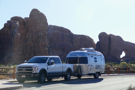 White truck with Airstream RV rental by red rock formations & an arch. Sunny desert road trip scene.