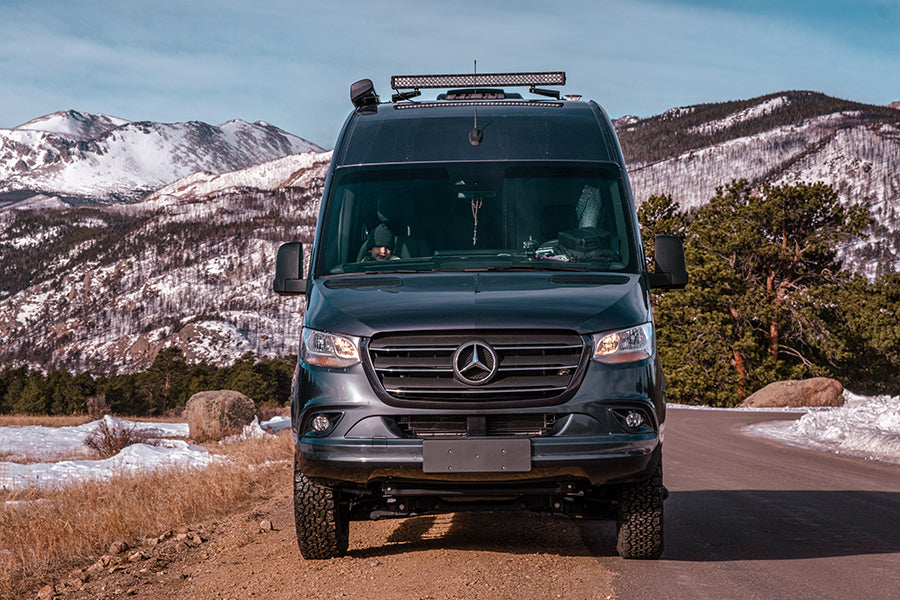Front view of a dark Mercedes Sprinter camper van, an excellent RV rental for mountain road trips amidst snowy peaks.