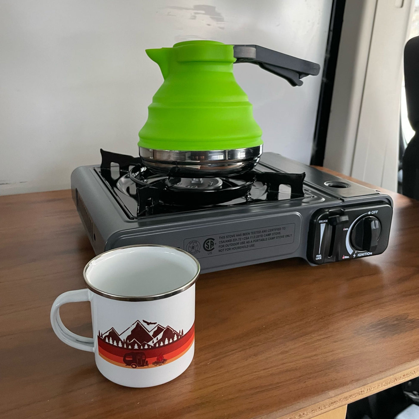 RV rental cooking: A bright green collapsible kettle sits on a portable camping stove next to a campfire-themed mug.