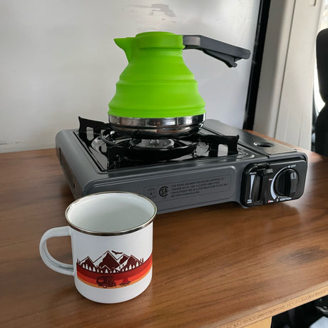 RV rental cooking: A bright green collapsible kettle sits on a portable camping stove next to a campfire-themed mug.