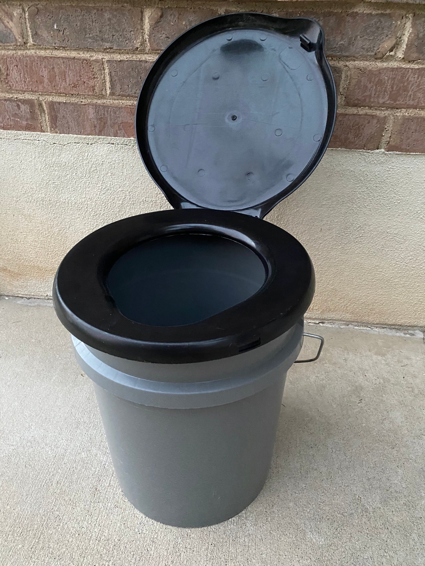A gray bucket-style portable toilet with a black seat and open black lid, perfect for your rv rental adventure.