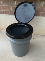 A gray bucket-style portable toilet with a black seat and open black lid, perfect for your rv rental adventure.