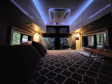 Inside a comfy RV rental: bed with patterned comforter, blue ambient lighting, and large windows revealing a peaceful forest.