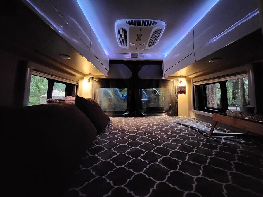 Inside a comfy RV rental: bed with patterned comforter, blue ambient lighting, and large windows revealing a peaceful forest.