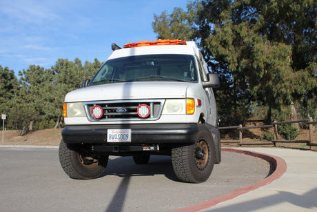 White custom adventure van, ideal for RV rental. Equipped for off-road travel, visible lifted suspension & recovery boards.