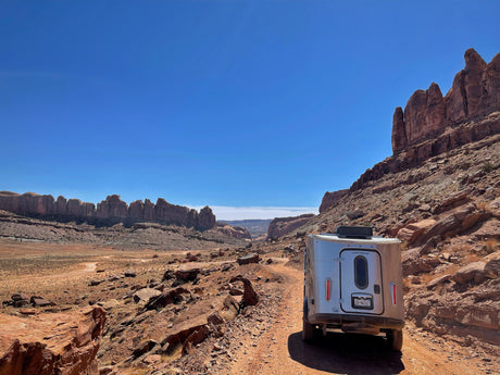 A silver Airstream rv rental on a rugged dirt road, surrounded by stunning desert rock formations under a clear blue sky.
