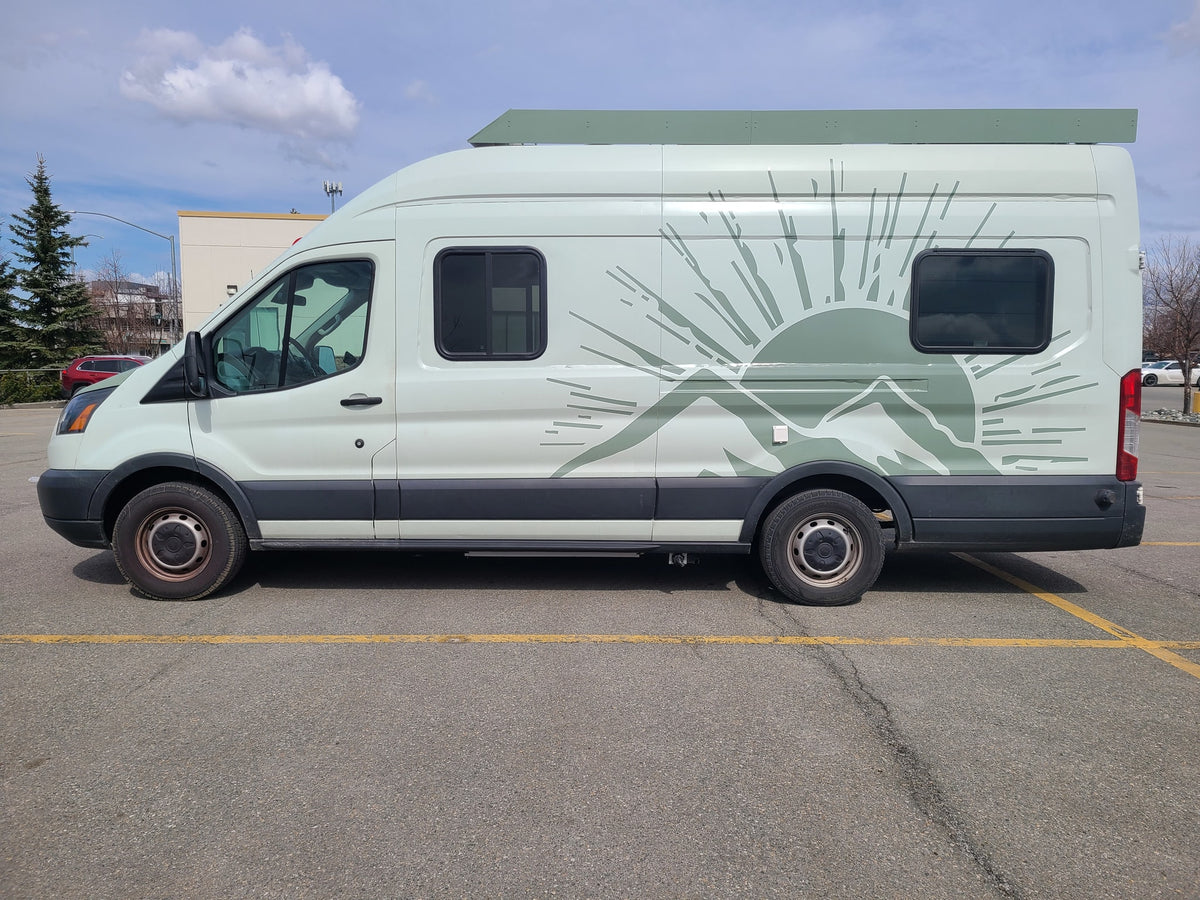 Light green RV rental camper van with mountain/sun graphic parked on asphalt under a partially cloudy sky.