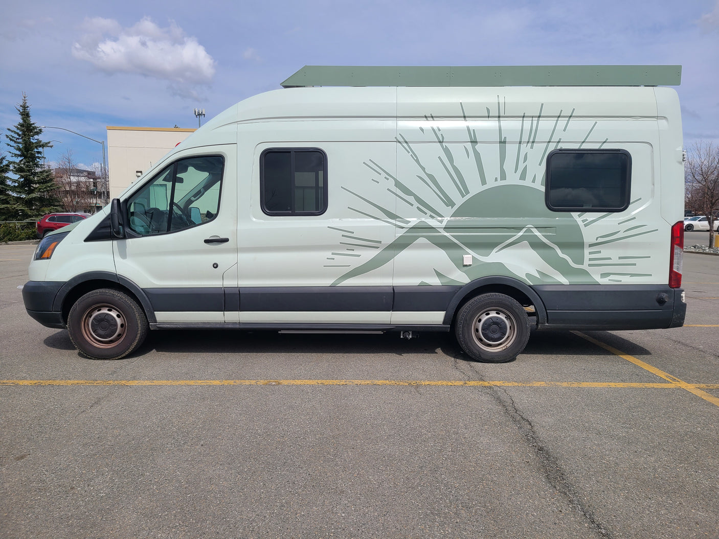 Light green RV rental camper van with mountain/sun graphic parked on asphalt under a partially cloudy sky.