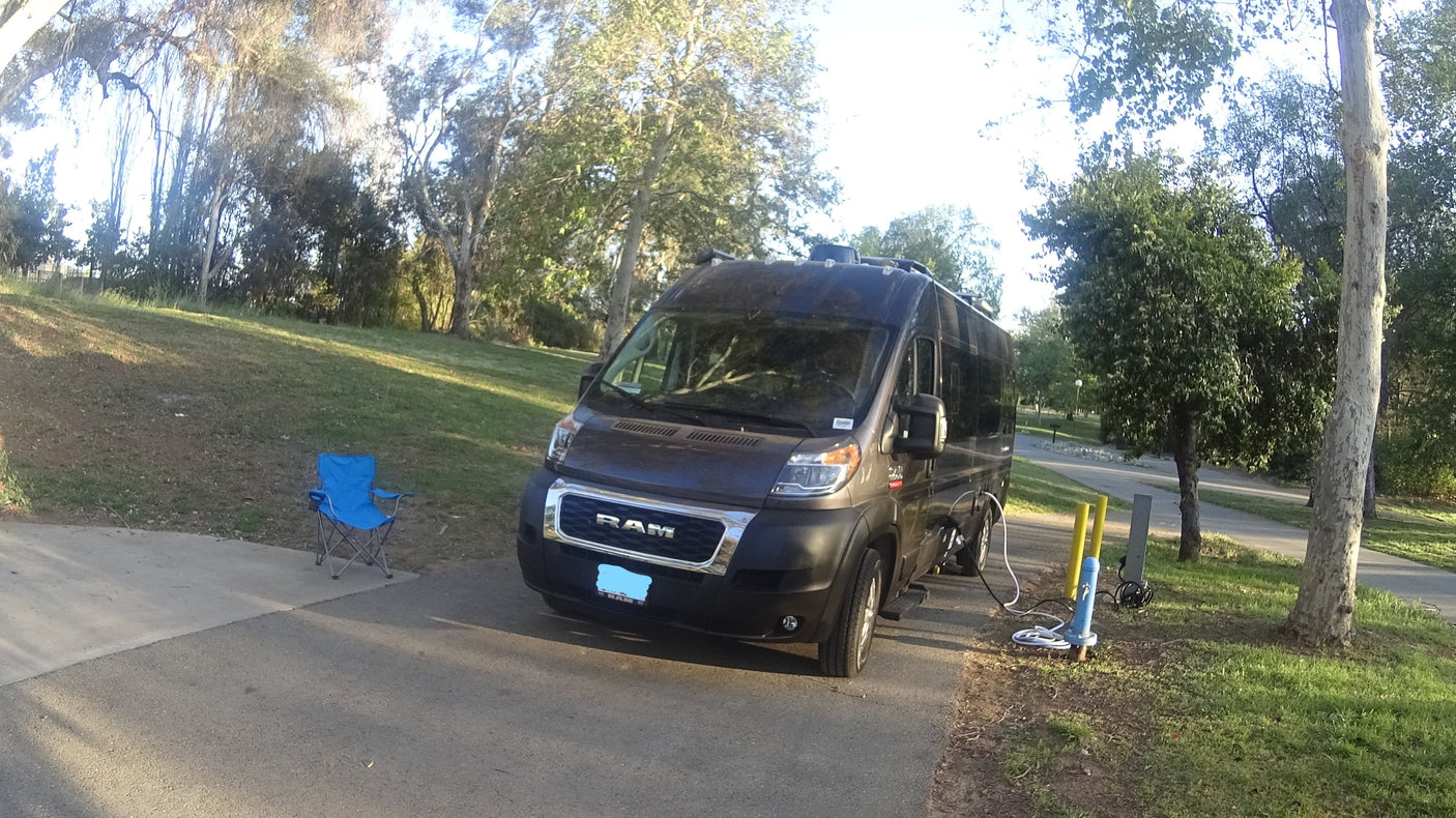 Dark gray RV rental camper van set up at a grassy park site with a blue chair and utility hookups.