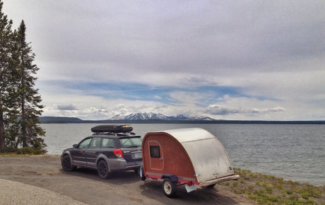 Subaru + teardrop RV rental parked lakeside, framed by pines and snow-capped mountains. Perfect for camping adventures!