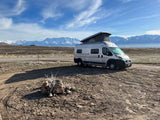 Scenic RV rental view: Camper van by a lake with snow-capped mountains under a blue sky.