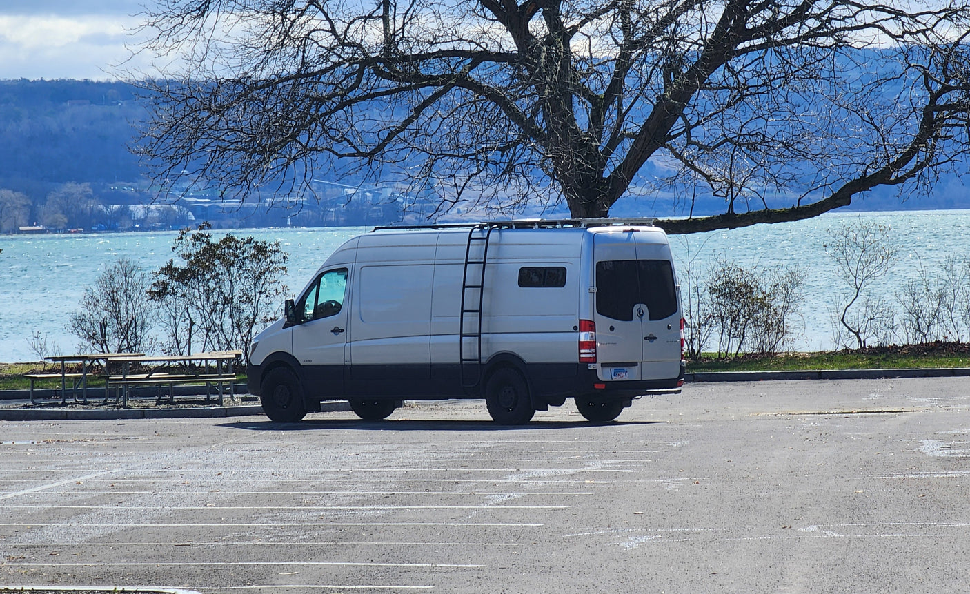 White Sprinter camper van (perfect for an RV rental) parked by a windy lake with bare trees and distant hills visible.