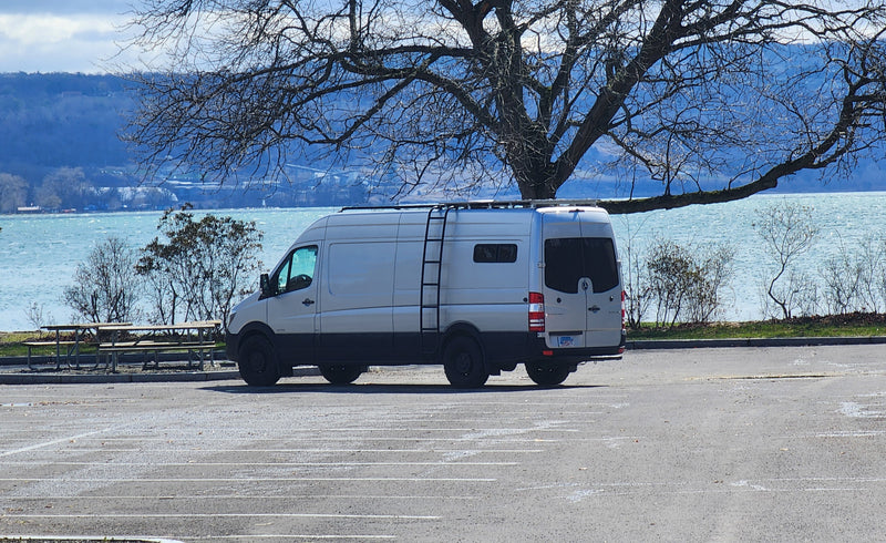White Sprinter camper van (perfect for an RV rental) parked by a windy lake with bare trees and distant hills visible. — photo 2