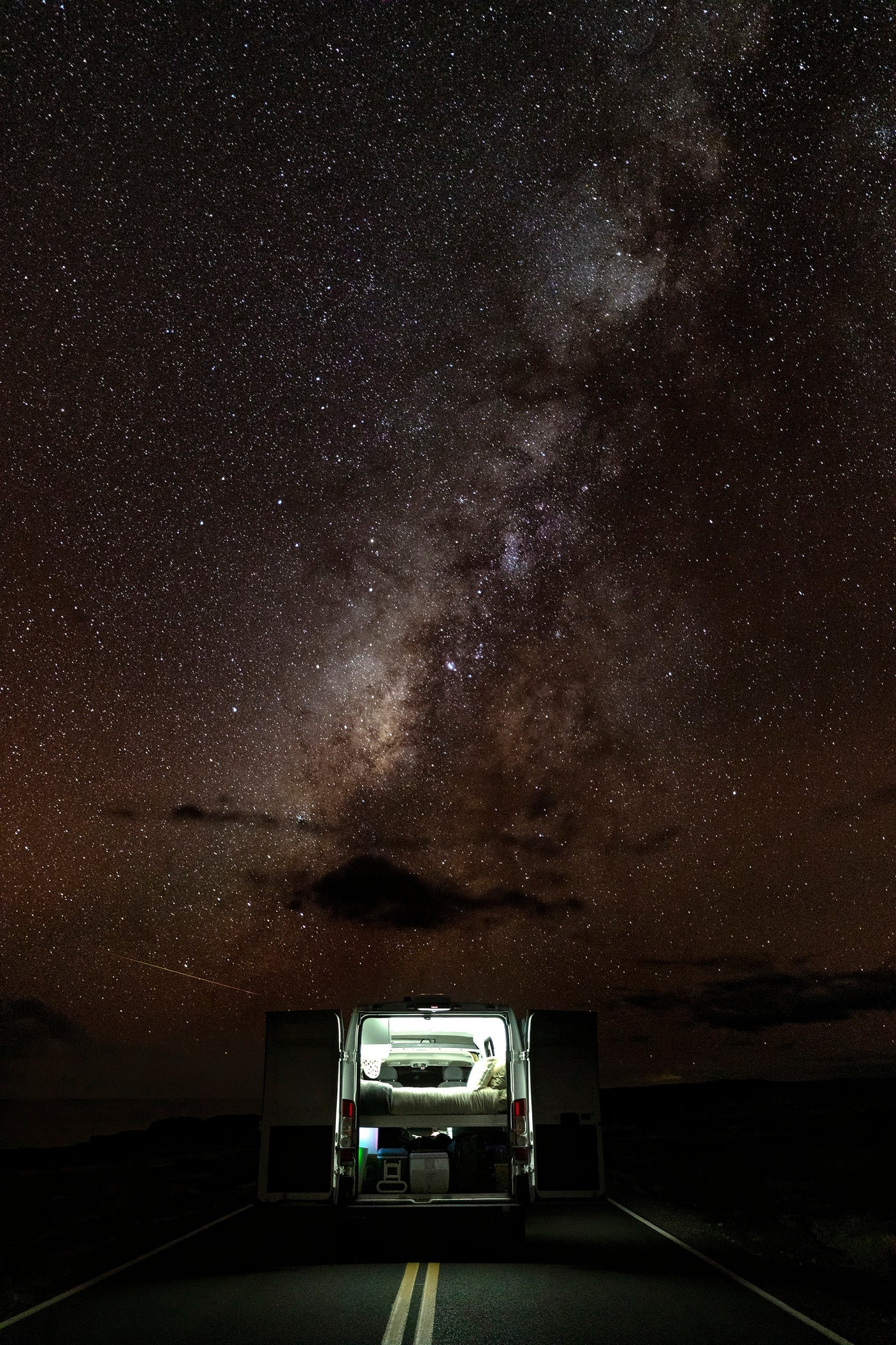 Experience an RV rental under the majestic Milky Way! A camper van with open back doors and illuminated interior, parked on a dark road.
