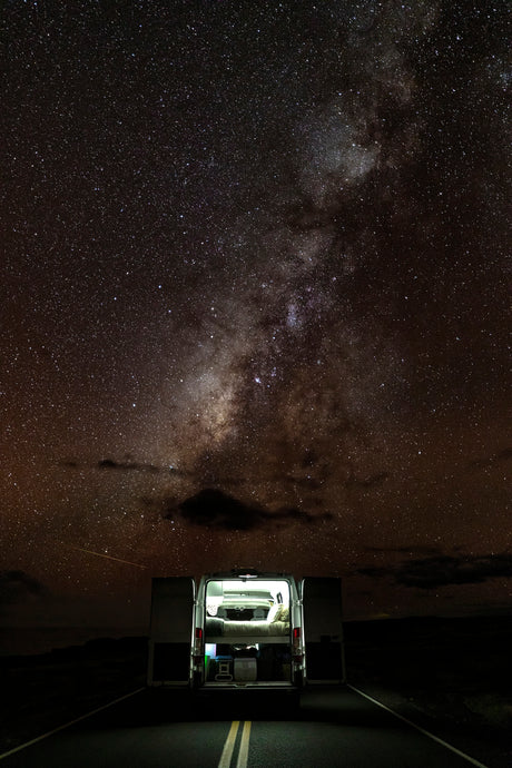 Experience an RV rental under the majestic Milky Way! A camper van with open back doors and illuminated interior, parked on a dark road.
