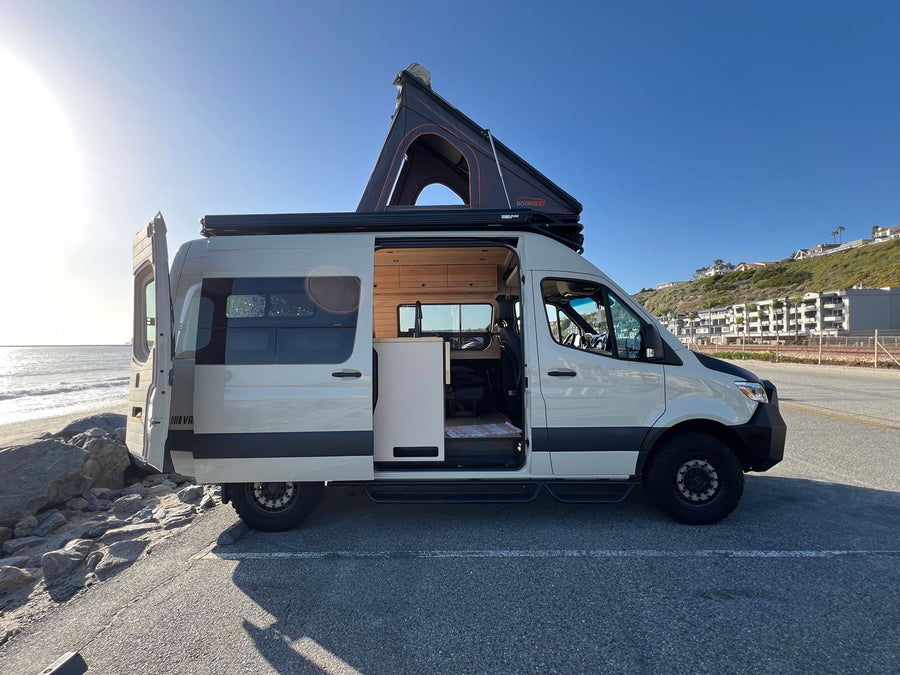 White RV rental camper van parked oceanfront with a deployed pop-top tent and open doors, ready for adventure.