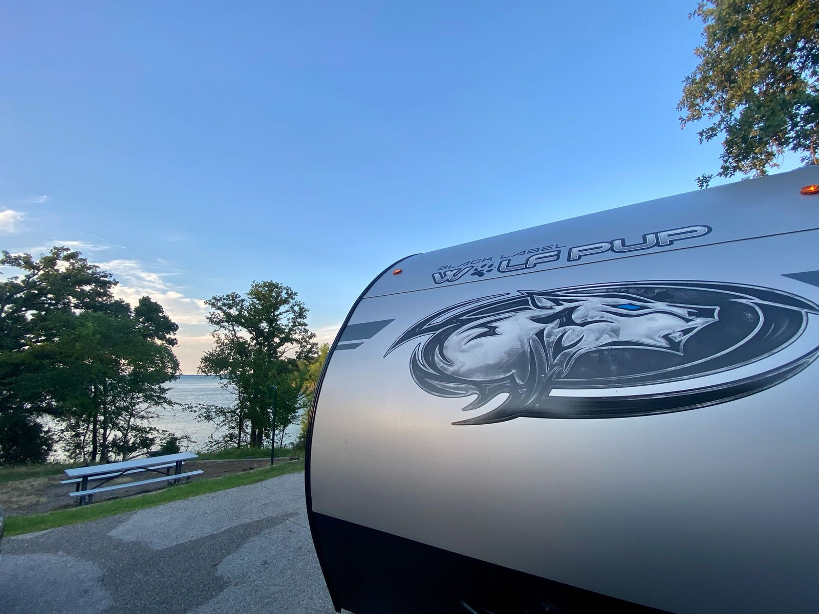 Lakeside bliss with our RV rental! The front of a 'Wolf Pup' travel trailer, parked by water, trees & a picnic table.