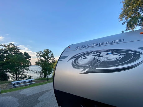 Lakeside bliss with our RV rental! The front of a 'Wolf Pup' travel trailer, parked by water, trees & a picnic table.