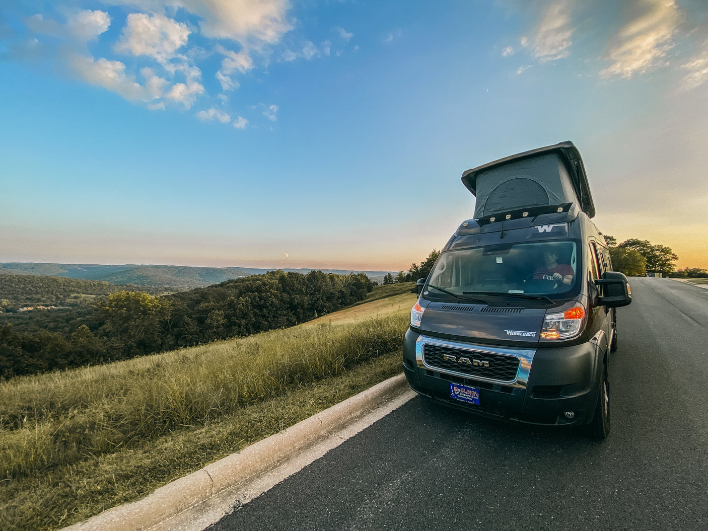 A Winnebago camper van RV rental with a pop-top, parked roadside overlooking a green valley under a sky with a crescent moon.