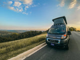 A Winnebago camper van RV rental with a pop-top, parked roadside overlooking a green valley under a sky with a crescent moon.