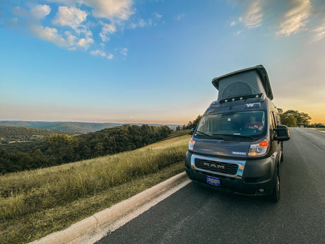 A Winnebago camper van RV rental with a pop-top, parked roadside overlooking a green valley under a sky with a crescent moon.