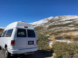 White RV rental parked with its back to the camera, facing snow-dusted mountains under a clear blue sky. Ideal for a Colorado adventure!