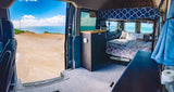 Spacious RV rental campervan interior with bed, kitchen, & an open door revealing a stunning ocean view.