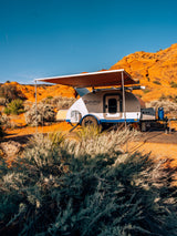 Pindrop teardrop RV rental with awning extended in a vibrant desert campsite, featuring orange rock formations.