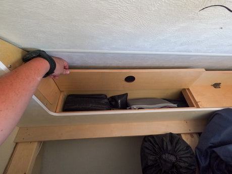 Checking overhead storage in an RV rental. A hand with a watch reaches into the wooden compartment holding travel bags and gear.