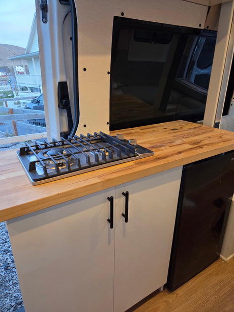 RV rental kitchen: gas stove, wood counter, white cabinets, mini-fridge. Outside view visible through open van door.