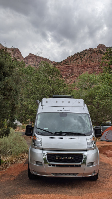 A silver RAM Roadtrek RV rental is parked at a scenic outdoor campsite with red rock mountains and green trees under grey skies.