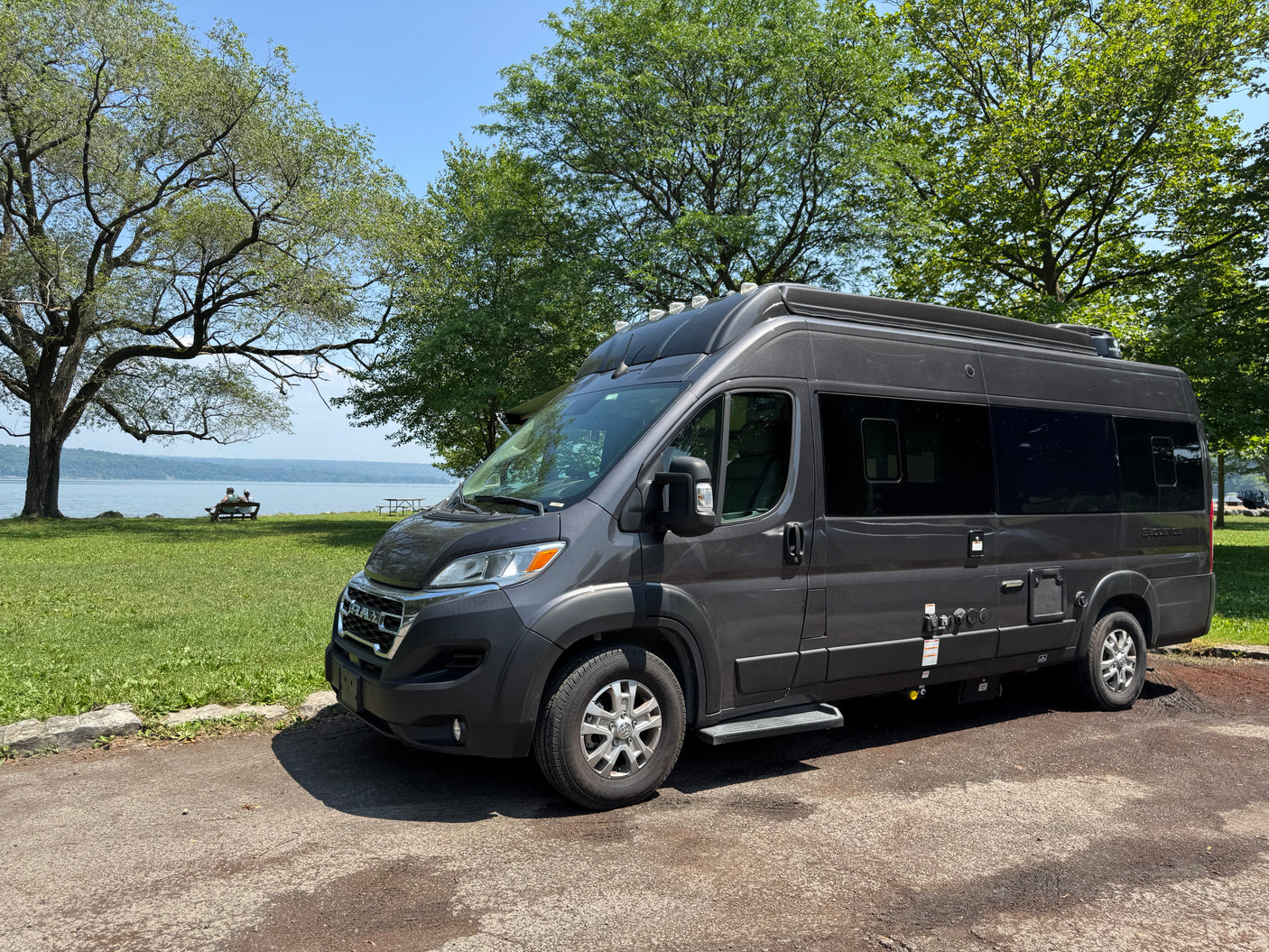 Scenic dark grey rv rental camper van parked by a serene lake with trees. Two people relax on a bench by the water.
