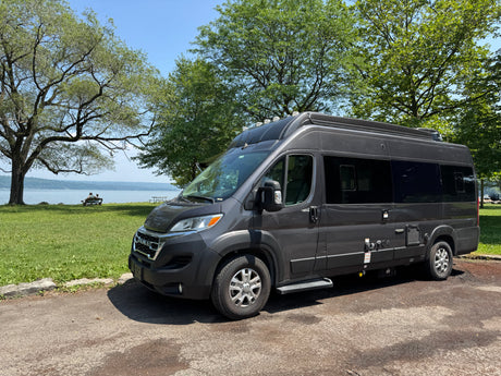 Scenic dark grey rv rental camper van parked by a serene lake with trees. Two people relax on a bench by the water.