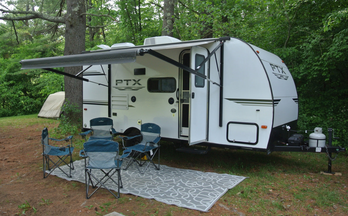 White travel trailer RV rental parked at a shady campsite. Awning extended over chairs and rug, ready for outdoor living.