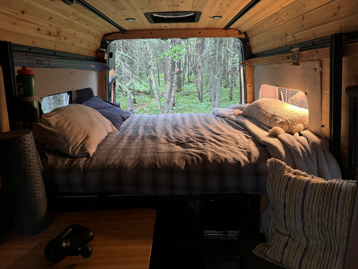 Relax in a cozy RV rental bed, surrounded by warm wood and a serene forest view out the open back.