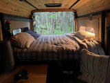 Relax in a cozy RV rental bed, surrounded by warm wood and a serene forest view out the open back.