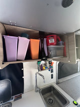 Inside an RV rental: a compact kitchen area with a sink, faucet, and colorful overhead storage bins.