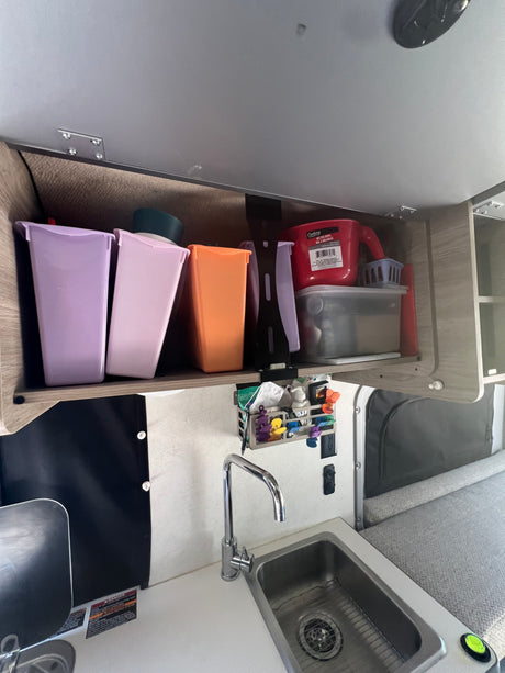 Inside an RV rental: a compact kitchen area with a sink, faucet, and colorful overhead storage bins.