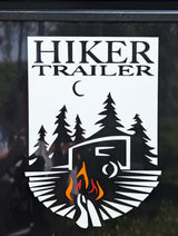 A white HIKER TRAILER RV rental emblem features a camper, pine trees, moon, and campfire on a black background.