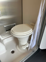 Inside an RV rental: a compact wet bath with white toilet, shower pan, drain, and a hanging white curtain.