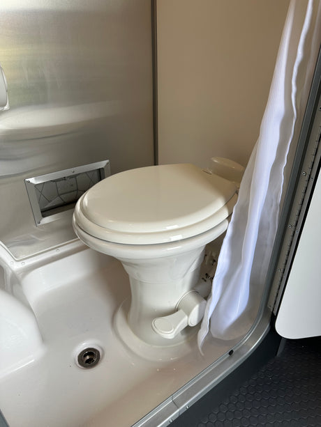 Inside an RV rental: a compact wet bath with white toilet, shower pan, drain, and a hanging white curtain.