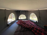 Inside an RV rental's rooftop tent: a cozy bed with a plaid blanket and arched windows offering scenic forest views. Ideal for camping.